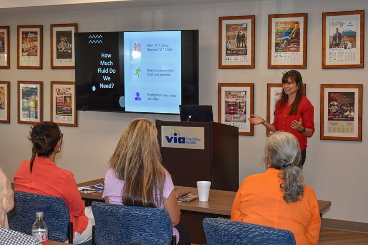 A woman stands at the front of a room in front of a group of people while delivering a presentation.