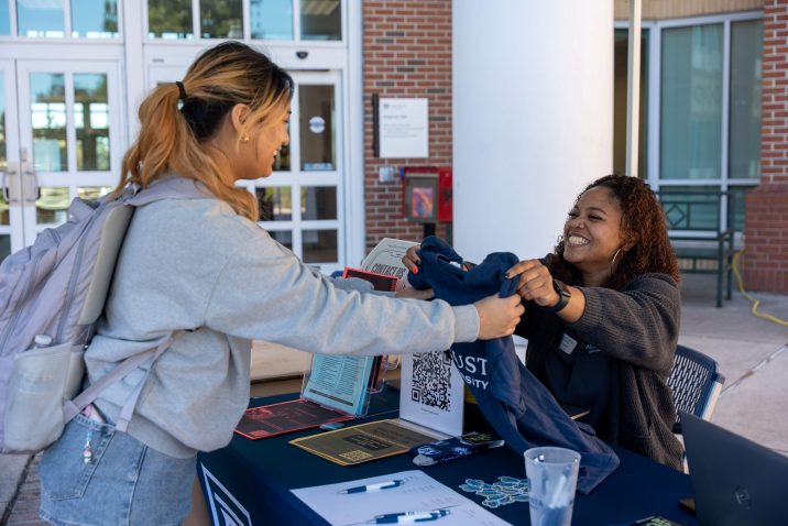 A woman hands another woman a t-shirt at a table outside during a special event.