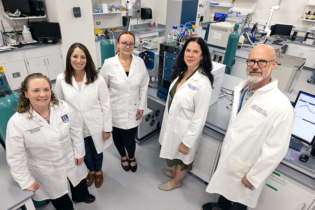A group of university faculty researchers in lab coats stand in a laboratory and pose for the camera.